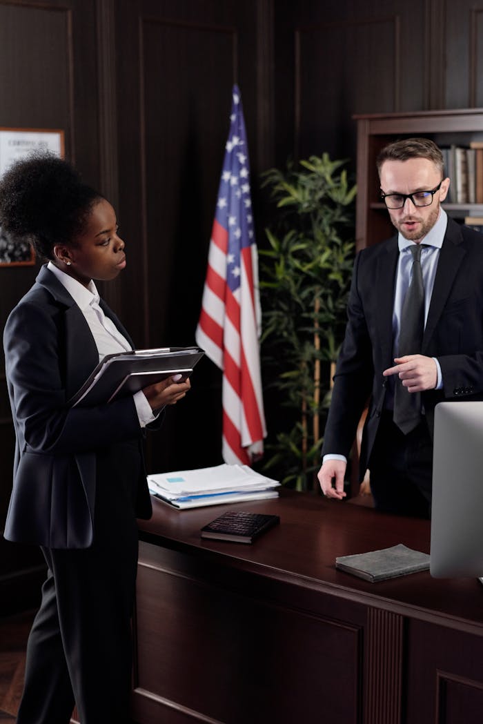 Two colleagues engaged in a discussion in a formal office setting with an American flag.